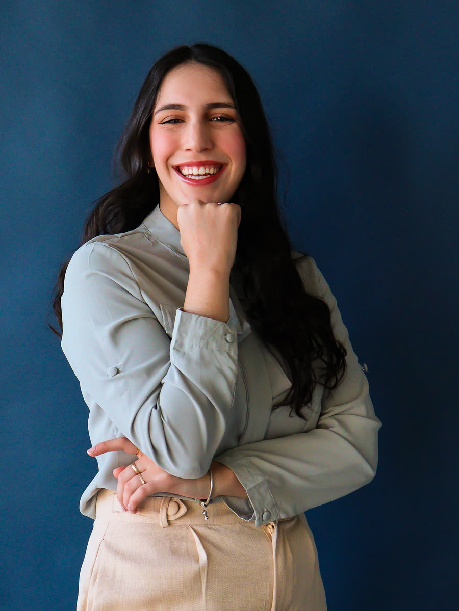 Dra. Manuelle Bianchi sorrindo, de camisa clara, em frente a um fundo azul
